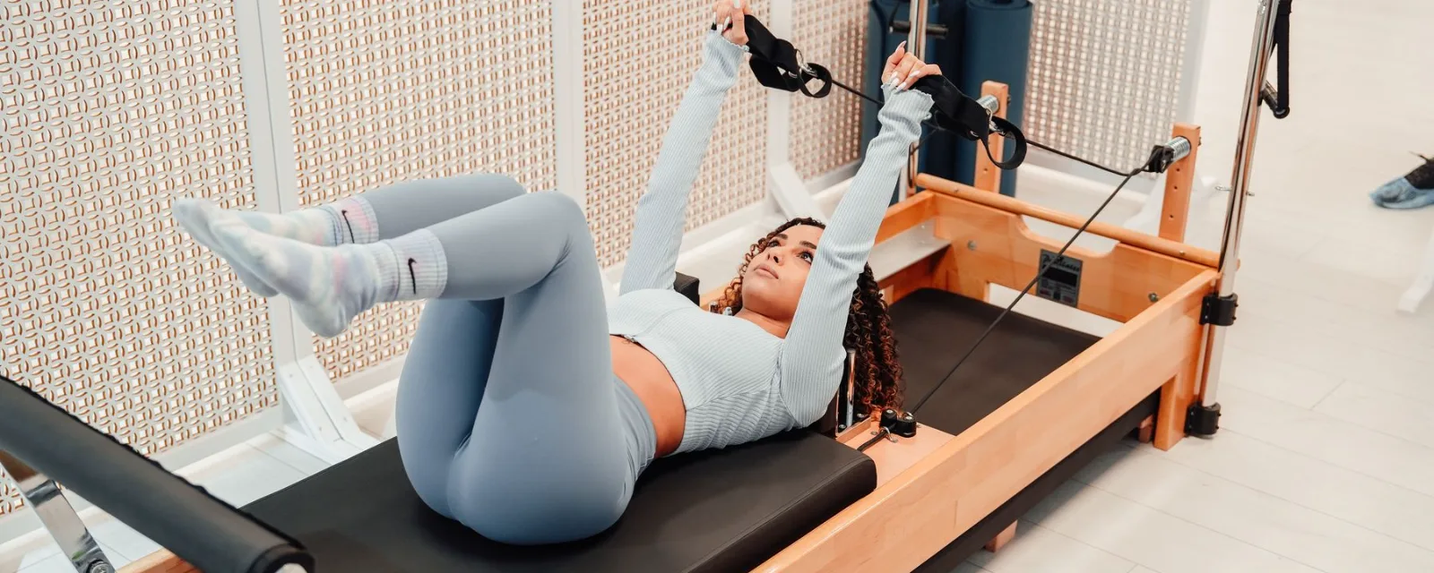 Une femme s'entra&icirc;ne sur un Pilates Reformer, allong&eacute;e sur le chariot, sangles en mains.